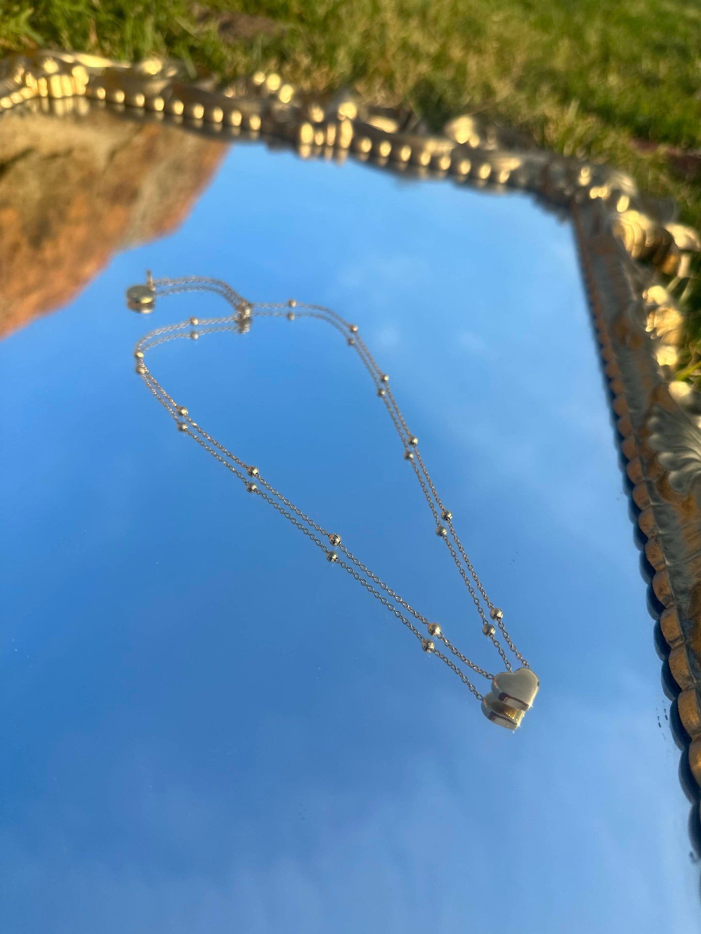 Gold chain necklace with a small gold heart-shaped pendant and small gold spheres evenly along the chain, with Shokhi Jewellery logo tag at the clasp, displayed on a mirror reflecting the sky