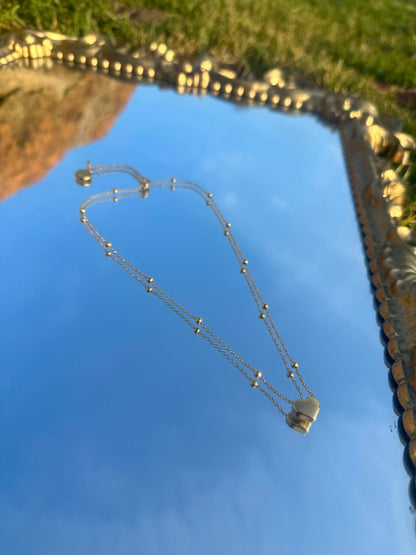 Gold chain necklace with a small gold heart-shaped pendant and small gold spheres evenly along the chain, with Shokhi Jewellery logo tag at the clasp, displayed on a mirror reflecting the sky