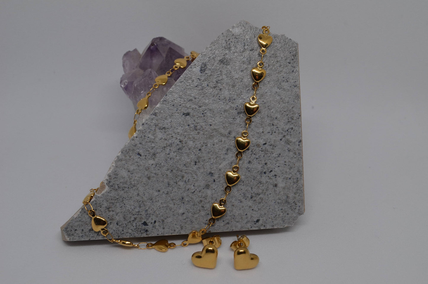 A gold heart-shaped chain necklace displayed with small gold heart-shaped earrings on an amethyst crystal slab