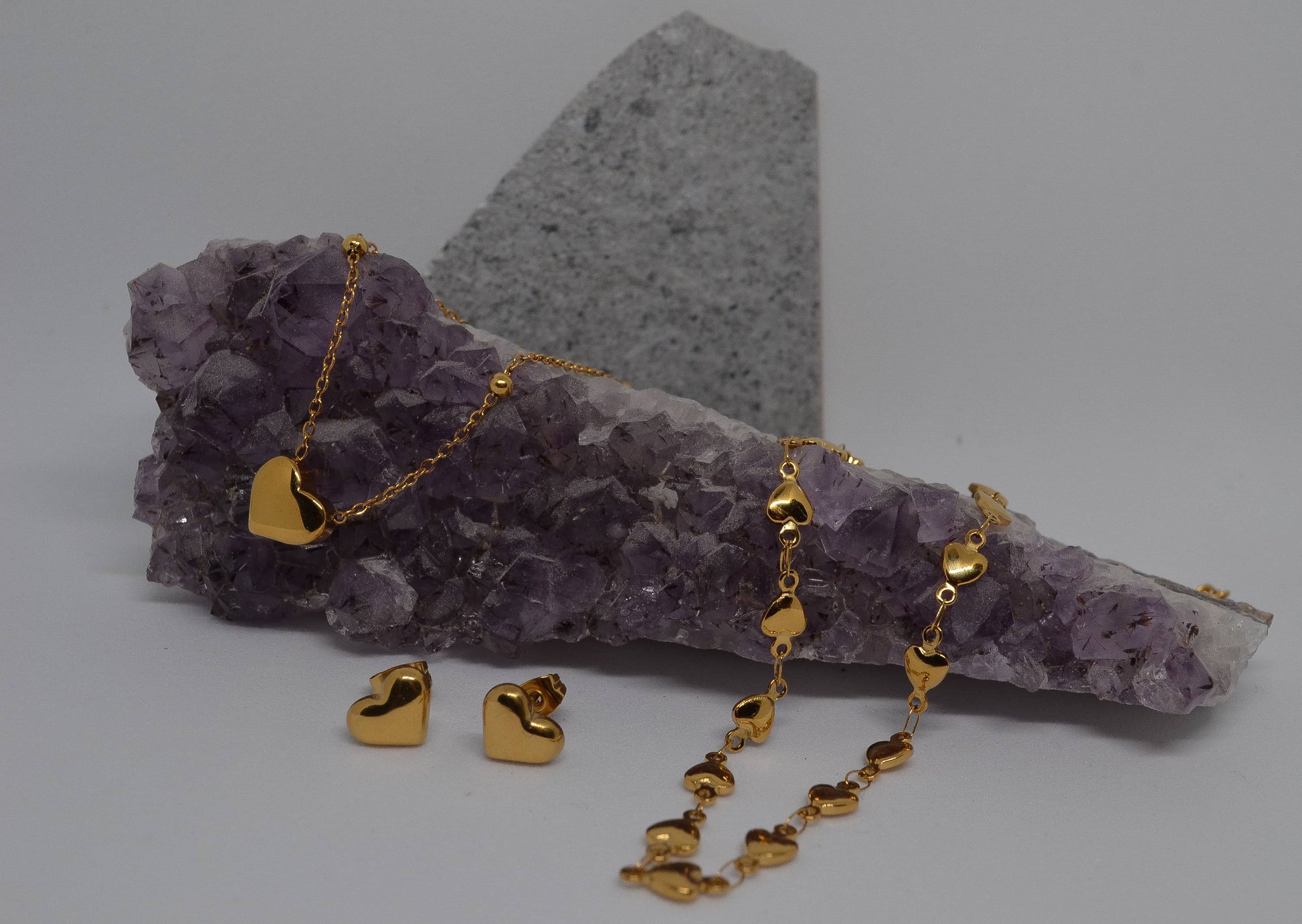 A gold heart-shaped chain necklace displayed with small gold heart-shaped earrings and a small gold heart-shaped pendant necklace on an amethyst crystal slab