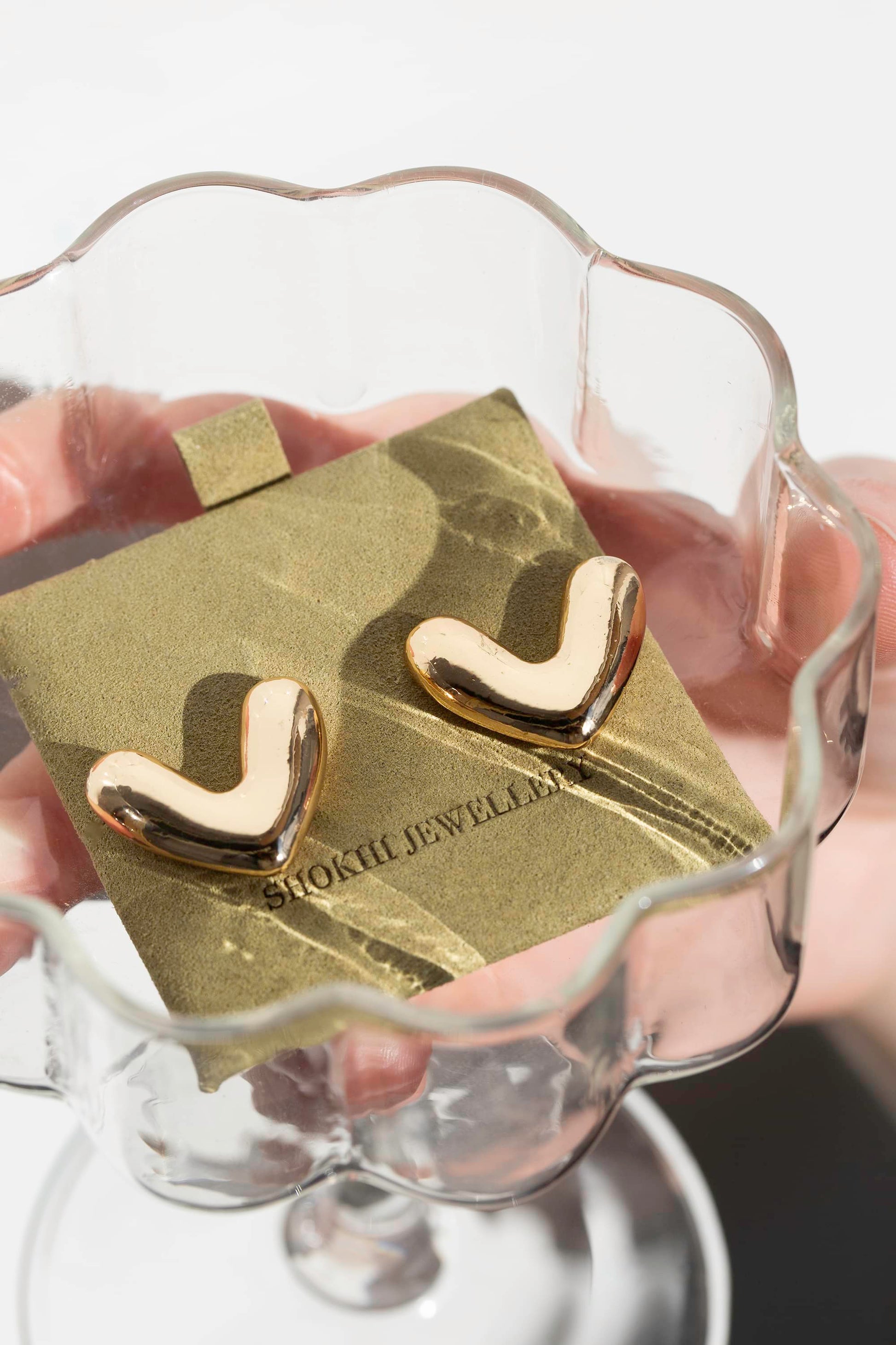 Thick heart-shaped gold stud earrings in their sage suede packaging in a cocktail glass