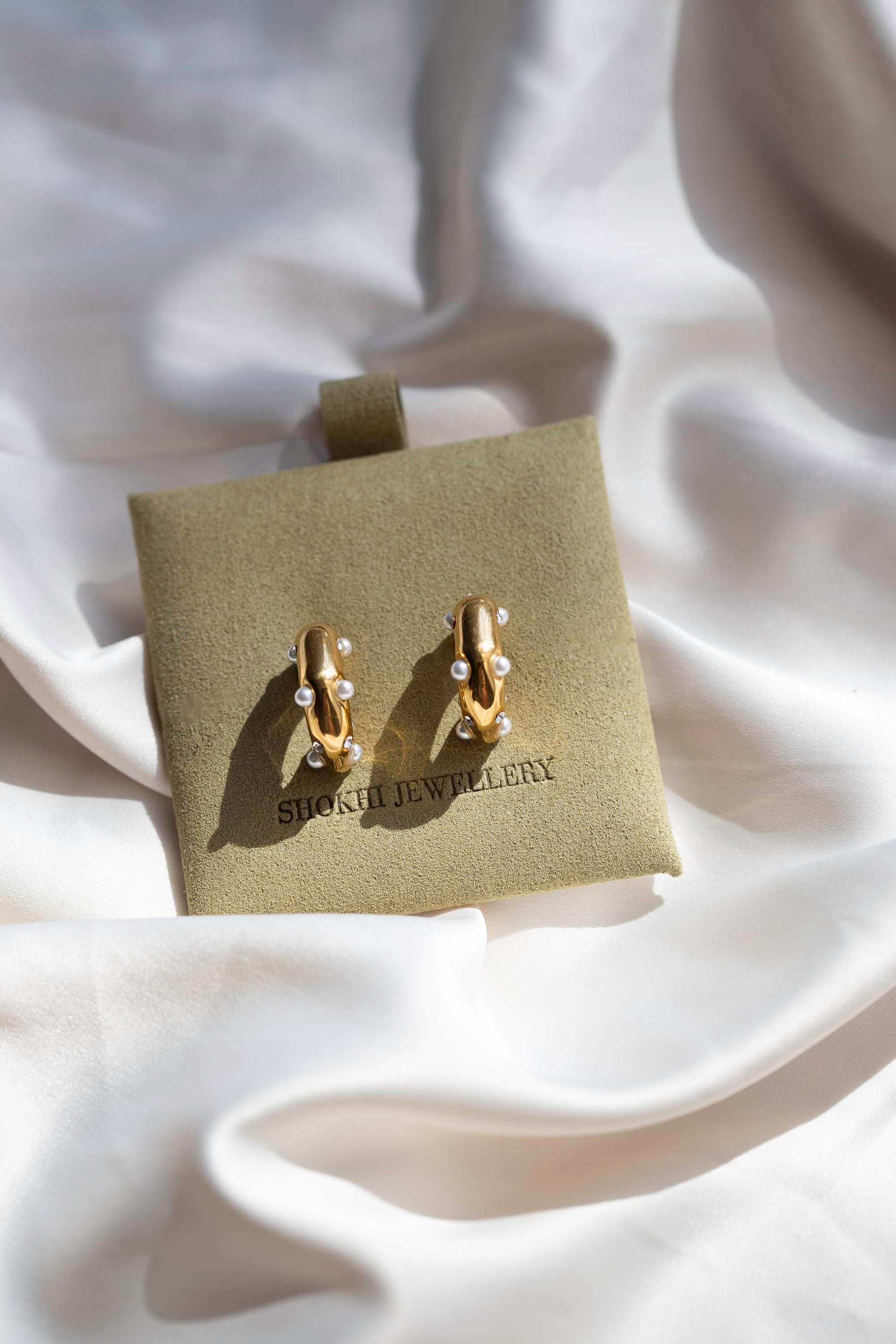 Gold stud earrings with a thick abstract design, featuring small pearl polka dots, displayed in sage suede packaging, on silk