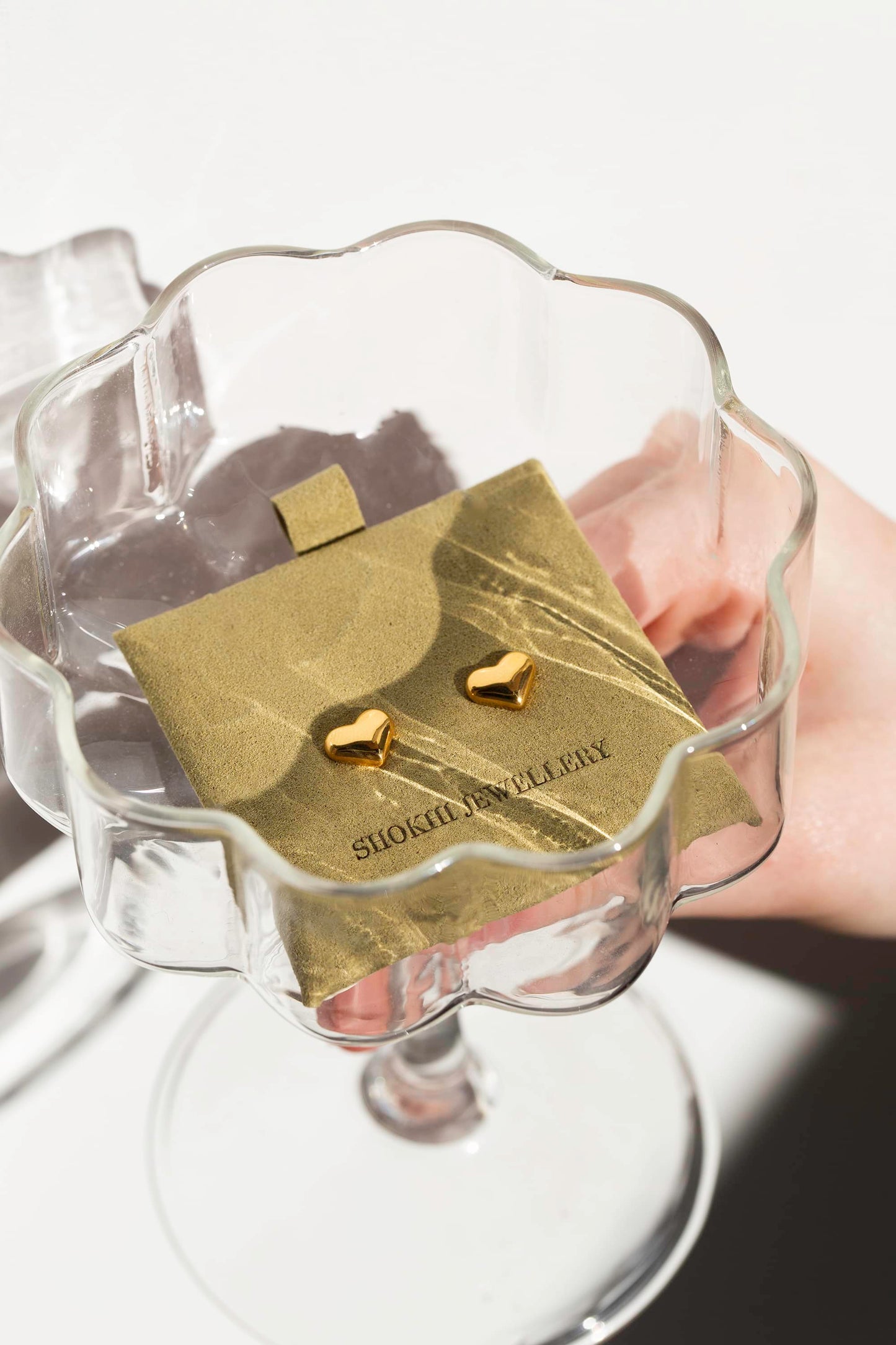 Small gold heart-shaped stud earrings on sage suede packaging in cocktail glass