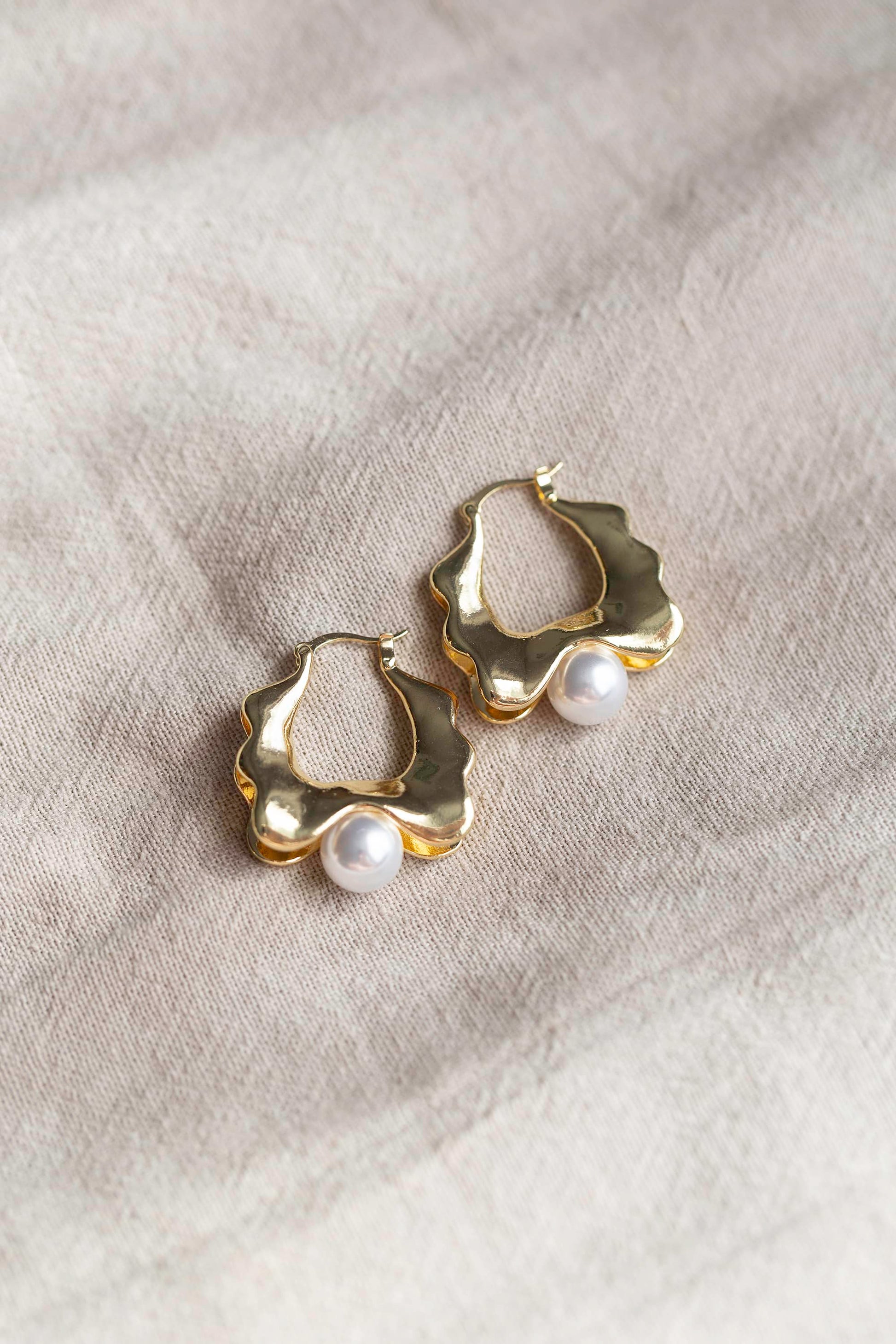 Close-up of gold hoop earrings with thick wavy edges and a large pearl at the bottom, displayed on linen