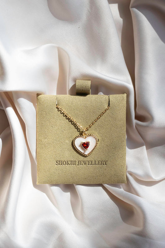 A gold and white heart-shaped pendant necklace with red heart-shaped gem set in the centre of the pendant, in sage suede packaging on silk