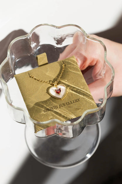 A gold and white heart-shaped pendant necklace with red heart-shaped gem set in the centre of the pendant, displayed in sage suede packaging in cocktail glass